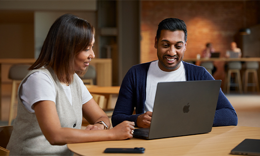 Amar collaborant avec quelqu’un tandis qu’il est assis avec son MacBook à une table dans une aire commune.
