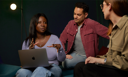 Ginelle, un MacBook sur les genoux, assise et travaillant avec deux collègues.