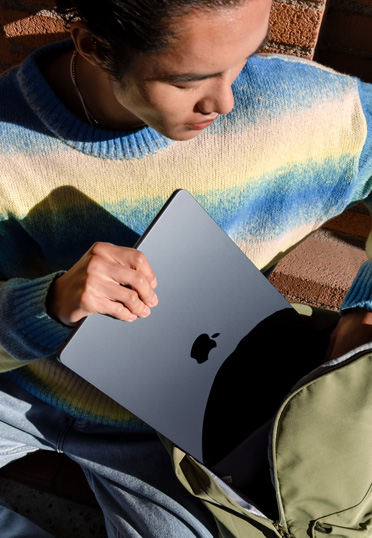 야외에 학생이 앉아 있는 모습. 닫힌 MacBook을 백팩에서 꺼내고 있습니다.
